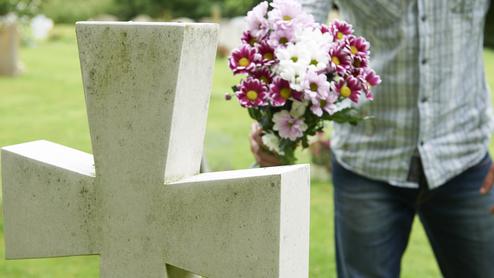 Mann mit einem Blumenstrauß steht neben einem Grabstein auf einem Friedhof in einer grünen Umgebung.